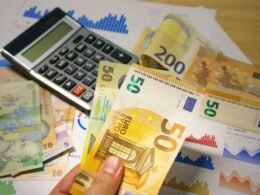 Eine Person hält einen Stapel Geldscheine in der einen Hand und einen Taschenrechner in der anderen auf einem Tisch.