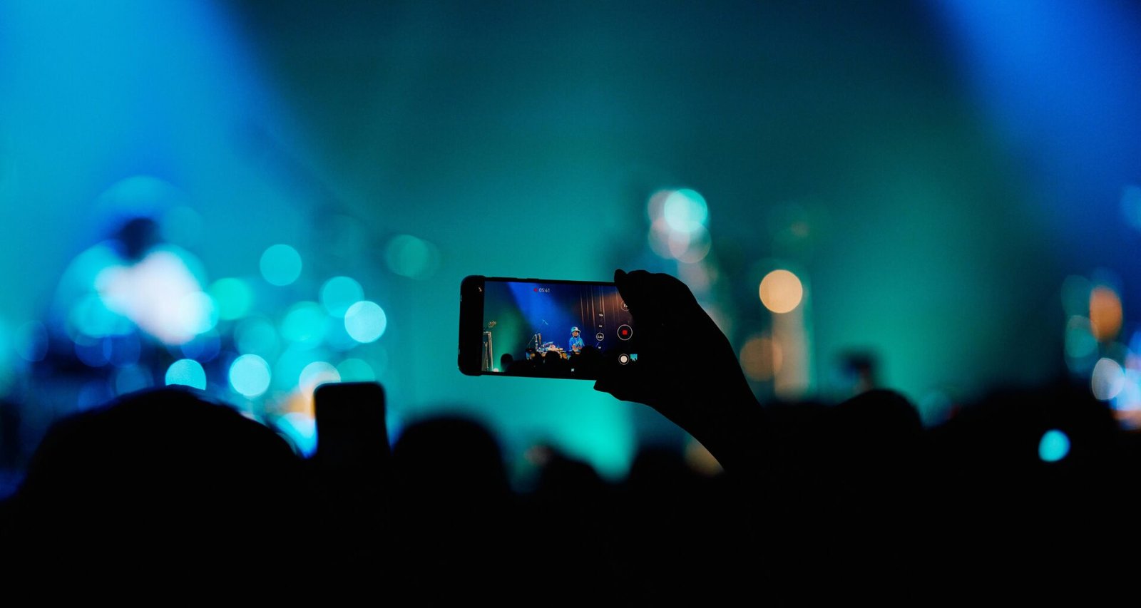Person fotografiert ein Konzert mit dem Smartphone inmitten einer Menschenmenge, Bühne und Lichter im Hintergrund.