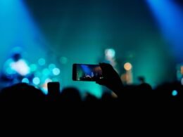 Person fotografiert ein Konzert mit dem Smartphone inmitten einer Menschenmenge, Bühne und Lichter im Hintergrund.