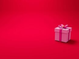 Ein pinker Geschenkkarton mit dekorativer Schleife auf rotem Hintergrund liegt zentral im Bild