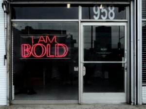 Rotes Neon-Leuchtschild mit der Aufschrift „I am bold“ an einer dunklen Wand, fotografiert von Max Böhme