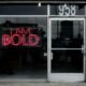 Rotes Neon-Leuchtschild mit der Aufschrift „I am bold“ an einer dunklen Wand, fotografiert von Max Böhme