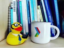 Eine weiße Kaffeetasse steht neben einem Stapel Bücher auf einem Holztisch, umgeben von natürlichem Licht.