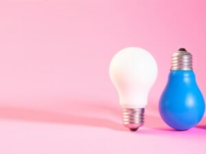 Eine weiße Glühbirne steht zwischen zwei blauen Glühbirnen auf hellem Untergrund. Foto von Sasun Bughdaryan.