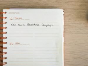 Notizbuch mit handschriftlicher Aufschrift ‚new year's resolution campaign‘ auf weißem Tisch, geeignet für Marketing-Artikel