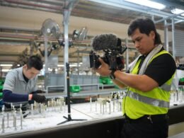 Ein Filmemacher filmt Mitarbeitende an einer automatisierten Produktionslinie in einer modernen Fabrikhalle