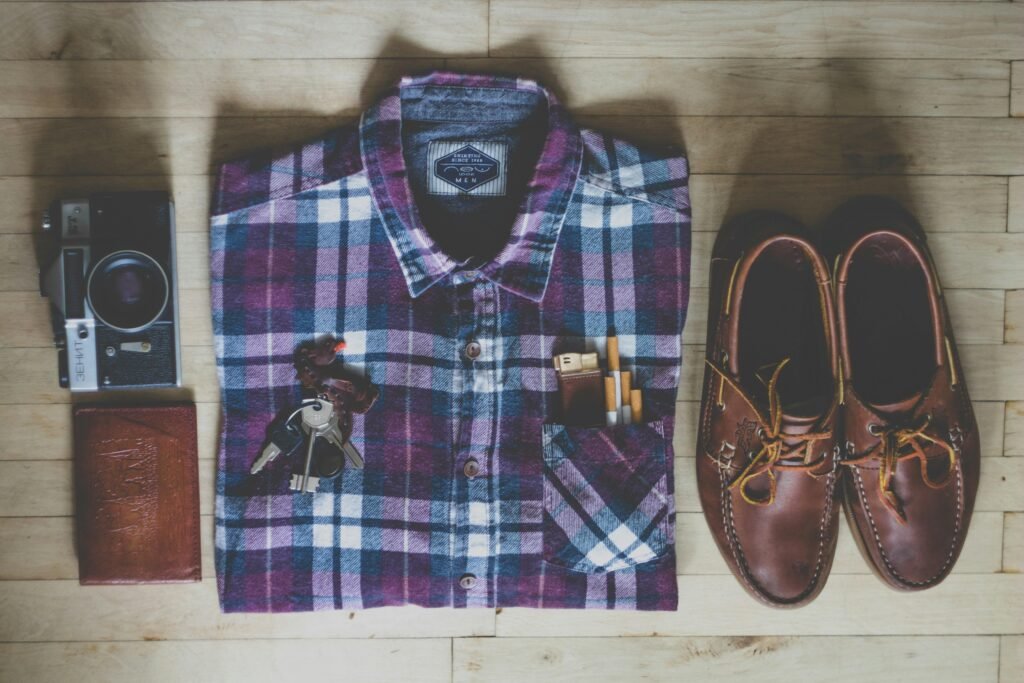 Flatlay mit Button-up-Hemd, Kamera, Schlüsseln, Zigaretten und braunen Leder-Bootsschuhen auf hellem Untergrund