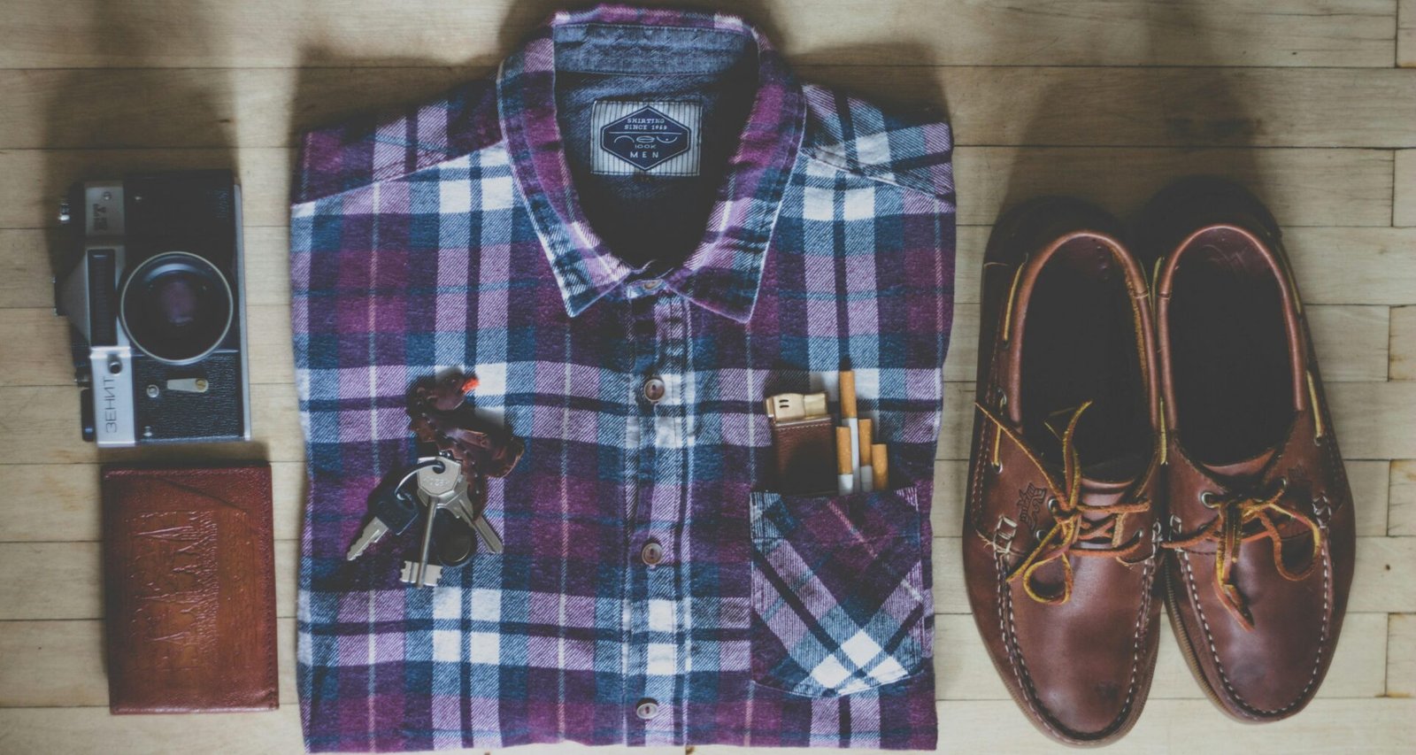 Flatlay mit Button-up-Hemd, Kamera, Schlüsseln, Zigaretten und braunen Leder-Bootsschuhen auf hellem Untergrund
