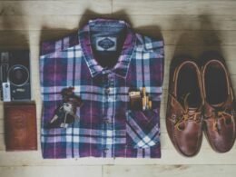 Flatlay mit Button-up-Hemd, Kamera, Schlüsseln, Zigaretten und braunen Leder-Bootsschuhen auf hellem Untergrund