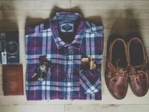 Flatlay mit Button-up-Hemd, Kamera, Schlüsseln, Zigaretten und braunen Leder-Bootsschuhen auf hellem Untergrund