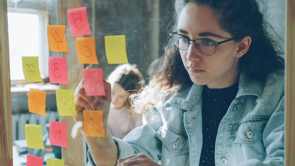 Frau betrachtet mehrere bunte Haftnotizen, die an einer Glaswand haften und Projekte strukturieren helfen.