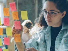 Frau betrachtet mehrere bunte Haftnotizen, die an einer Glaswand haften und Projekte strukturieren helfen.