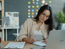 Frau sitzt am Schreibtisch und arbeitet mit Laptop und Notizbuch in einem modernen Arbeitsumfeld.