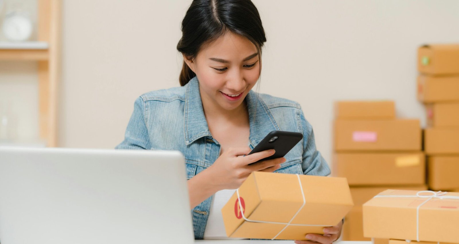 Frau prüft ein Paket mit dem Smartphone neben Laptop und Versandkartons im Homeoffice