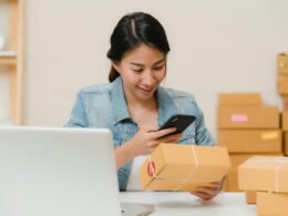 Frau prüft ein Paket mit dem Smartphone neben Laptop und Versandkartons im Homeoffice