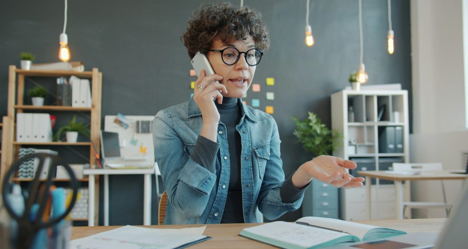 Frau sitzt an einem Schreibtisch im Büro und spricht am Telefon, moderne Arbeitsumgebung mit Computer im Hintergrund.