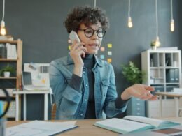 Frau sitzt an einem Schreibtisch im Büro und spricht am Telefon, moderne Arbeitsumgebung mit Computer im Hintergrund.