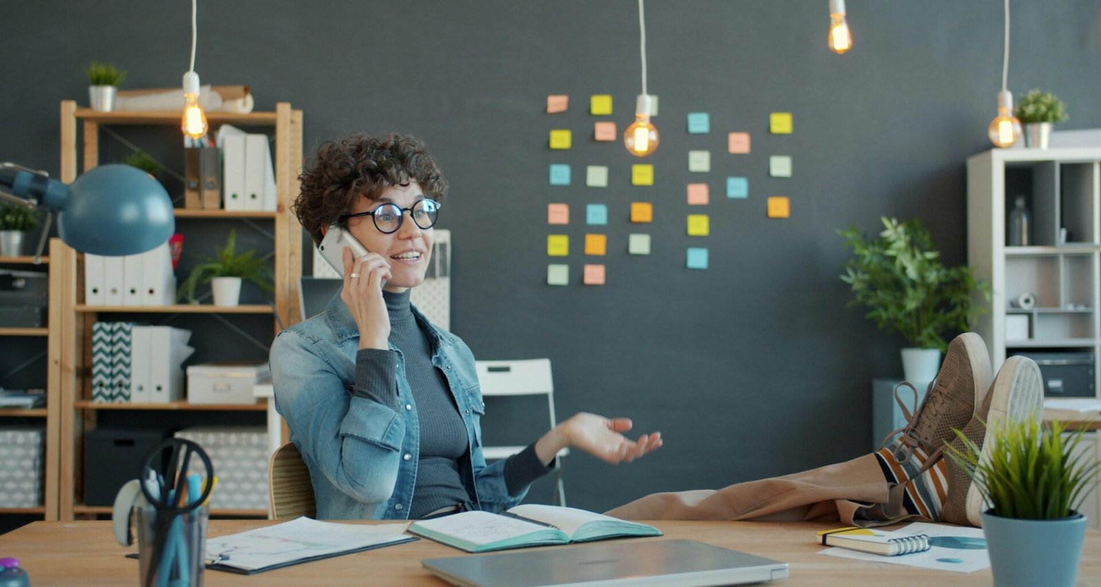 Frau sitzt mit hochgelegten Füßen am Schreibtisch und telefoniert im Homeoffice, moderner Arbeitsplatz mit Laptop und Notizbuch
