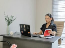 Frau arbeitet an einem Schreibtisch mit Laptop, umgeben von Notizbuch und Stift, in einem modernen Büro