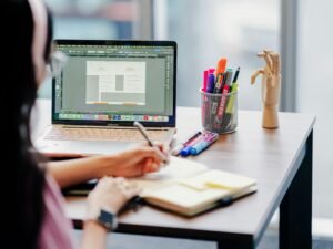 Frau sitzt an einem Arbeitsplatz und arbeitet konzentriert mit Laptop und Notizbuch an einem gut beleuchteten Schreibtisch.