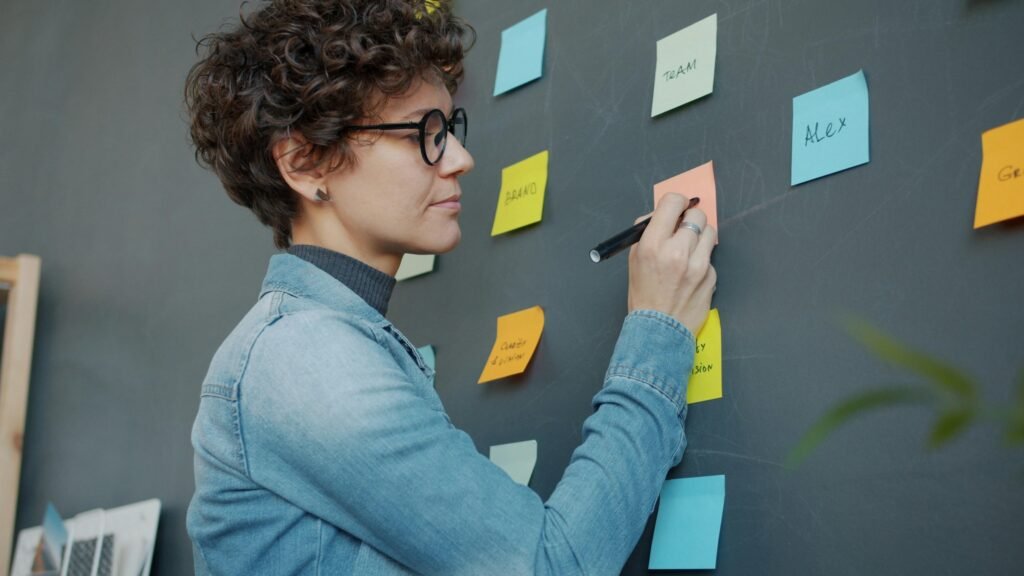 Frau schreibt auf bunte Haftnotizen an einer Wand, um Ideen im Team zu strukturieren und Arbeitsprozesse zu visualisieren