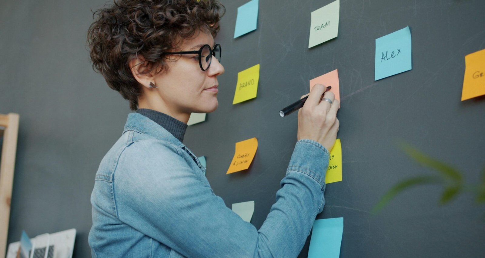 Frau schreibt auf bunte Haftnotizen an einer Wand, um Ideen im Team zu strukturieren und Arbeitsprozesse zu visualisieren