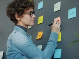 Frau schreibt auf farbige Haftnotizen an einer Wand, um Ideen und Aufgaben im Rahmen eines Projekts zu strukturieren.