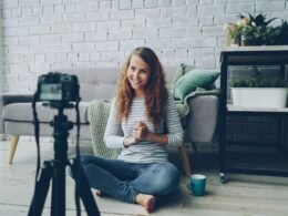 Frau sitzt auf dem Fußboden und filmt sich selbst mit einer Kamera; ideal illustriert für Artikel über Content-Erstellung