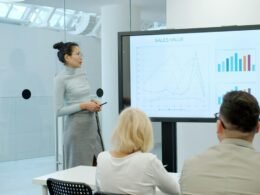 Eine Frau steht vor einer Gruppe und erklärt ein Balkendiagramm auf einem Bildschirm als Teil einer Präsentation.