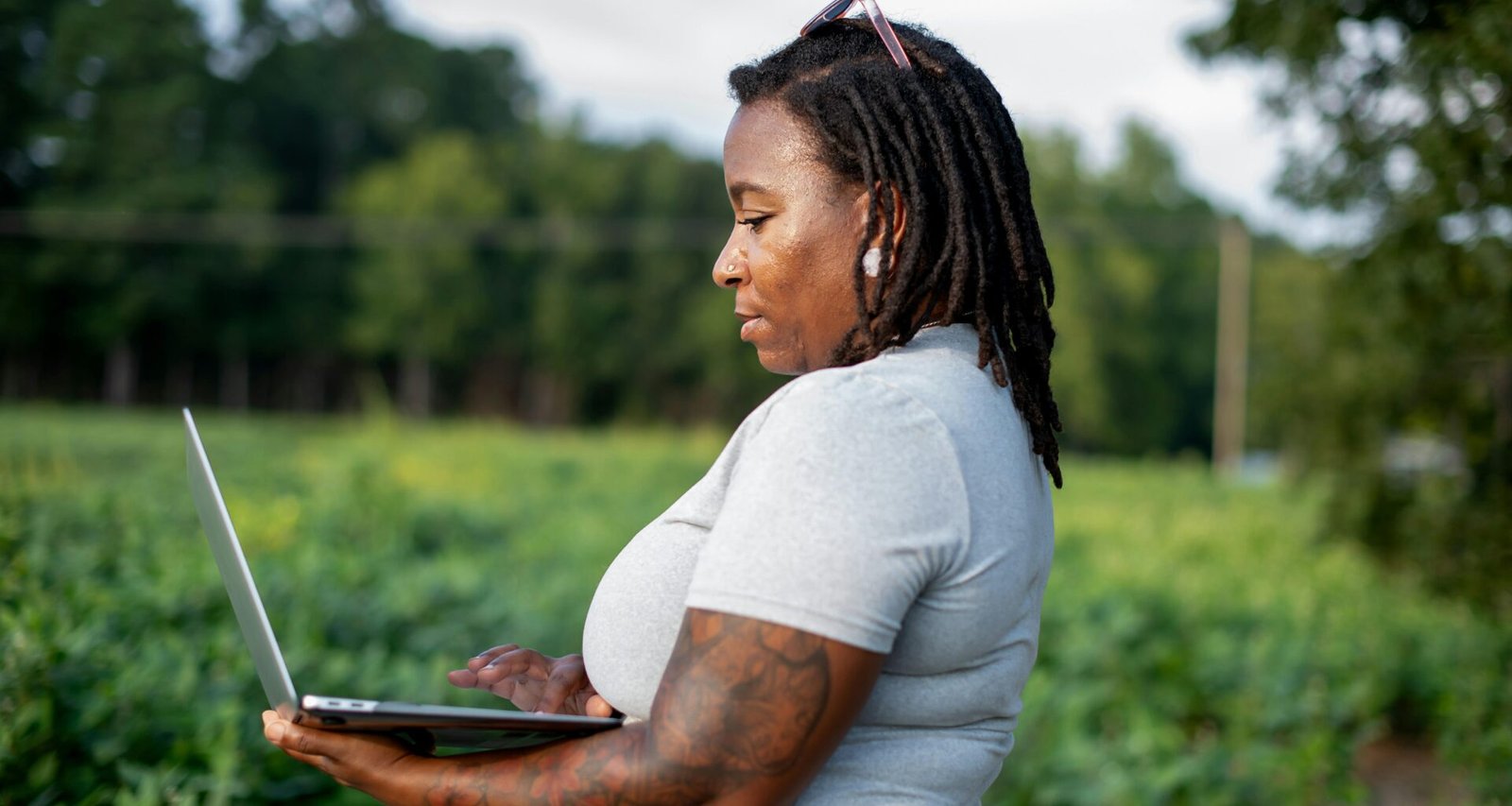 Frau sitzt mit Laptop auf dem Schoß in einem offenen Feld und arbeitet im Grünen an einem sonnigen Tag