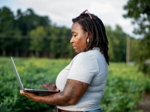 Frau sitzt mit Laptop auf dem Schoß in einem offenen Feld und arbeitet im Grünen an einem sonnigen Tag