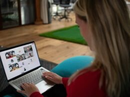 Frau sitzt am Schreibtisch und nimmt an einer Videokonferenz auf dem Laptop teil, mehrere Teilnehmer sind auf dem Bildschirm sichtbar.