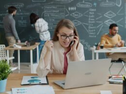 Frau sitzt am Schreibtisch, telefoniert und jubelt über einen beruflichen Erfolg während eines Business-Calls.