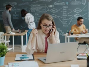 Frau sitzt am Schreibtisch, telefoniert und jubelt über einen beruflichen Erfolg während eines Business-Calls.