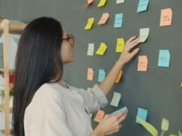 Frau platziert bunte Sticky Notes auf einer dunklen Wand als Teil eines kreativen Arbeitsprozesses im Büro.