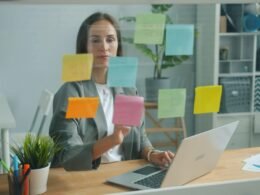 Frau ordnet bunte Haftnotizen auf einer Glaswand für Brainstorming und Projektplanung im modernen Büro.