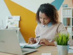 Frau sitzt an einem Schreibtisch und arbeitet mit Laptop und Notizbuch in einem modernen Büro