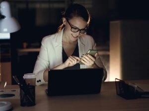 Frau arbeitet bei Nacht mit Laptop und Smartphone an einem Schreibtisch in einem dunklen Raum