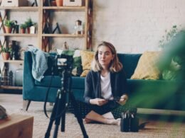 Eine Frau steht im Wohnzimmer und filmt sich selbst mit einer Digitalkamera auf einem Stativ neben dem Sofa.