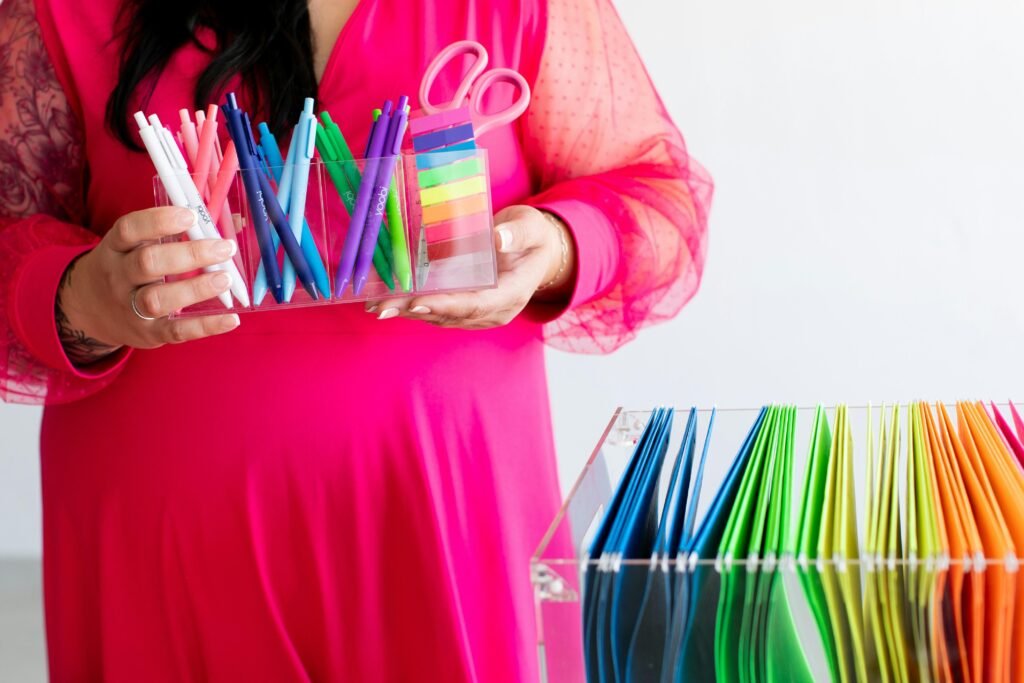 Frau in rosa Kleid hält mehrere bunte Stifte und eine Schere in der Hand vor neutralem Hintergrund