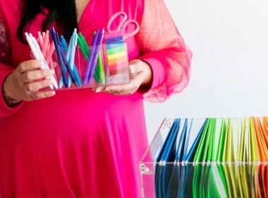 Frau im pinken Kleid hält bunte Stifte und eine Schere, Symbolbild für kreative Arbeitsumgebungen und Bastelthemen.