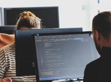 Frau in schwarzem Shirt sitzt am Schreibtisch neben einem großen, schwarzen Monitor in einem modernen Büro.