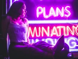 Frau mit weißem langen Shirt sitzt auf dem Fußboden in einem hellen Raum, Blick nach vorne gerichtet