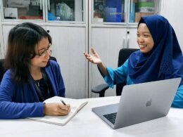 Frau in blauer Robe arbeitet konzentriert an einem MacBook an einem Tisch, geeignet für Themen wie Homeoffice und Technologie.