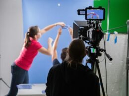 Eine Frau im pinken Langarmshirt hält eine schwarze Videokamera, Fokus auf technisches Interesse und Medieneinsatz.