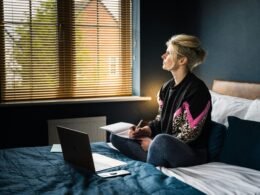 Frau in schwarzem und pinkem Blumenhemd sitzt mit Laptop auf dem Bett und arbeitet im Homeoffice