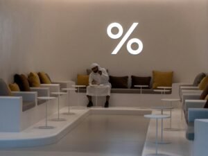 Frau mit weißem Langarmshirt und schwarzer Hose sitzt entspannt auf einem weißen Stuhl vor hellem Hintergrund