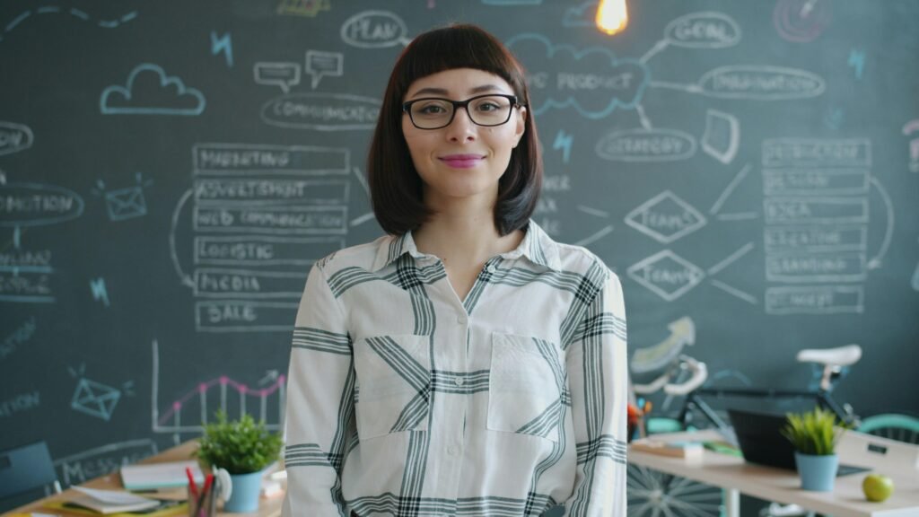 Frau mit Brille steht lächelnd vor einer Kreidetafel mit Aufzeichnungen im Hintergrund.