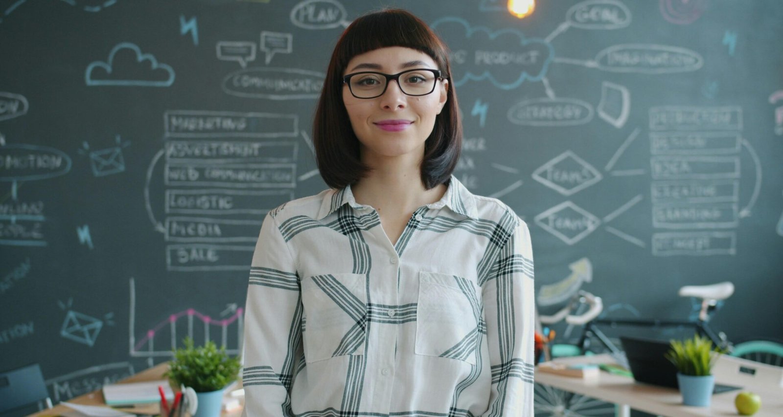 Frau mit Brille steht lächelnd vor einer Kreidetafel mit Aufzeichnungen im Hintergrund.