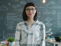 Frau mit Brille steht lächelnd vor einer Kreidetafel mit Aufzeichnungen im Hintergrund.